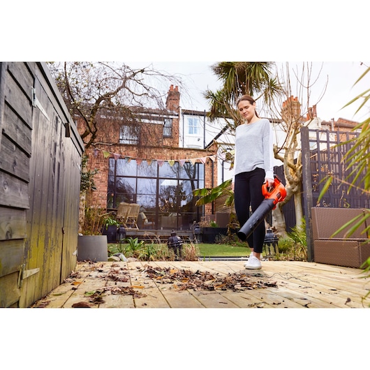 BLACK+DECKER BCBL200L25 leaf blower held by person clearing leaves on wooden deck in a garden.