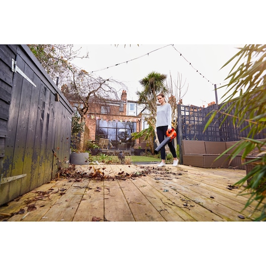 BLACKDECKER BCBL200L25 leaf blower clearing leaves on wooden deck in a garden setting.