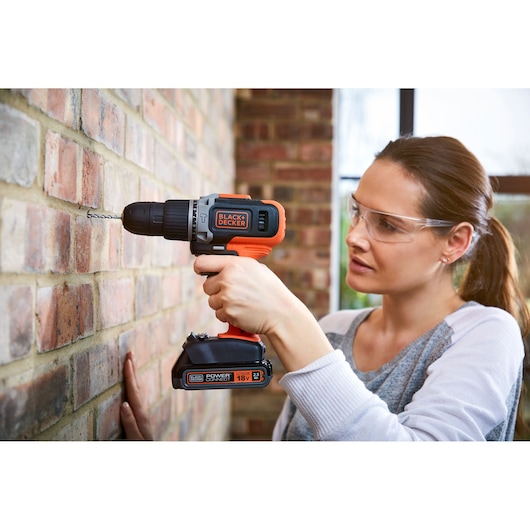 A person using a BLACK+DECKER cordless drill to make a hole in a brick wall. The drill is orange and black with visible branding and an 18V lithium battery. The person's face is blurred for privacy.