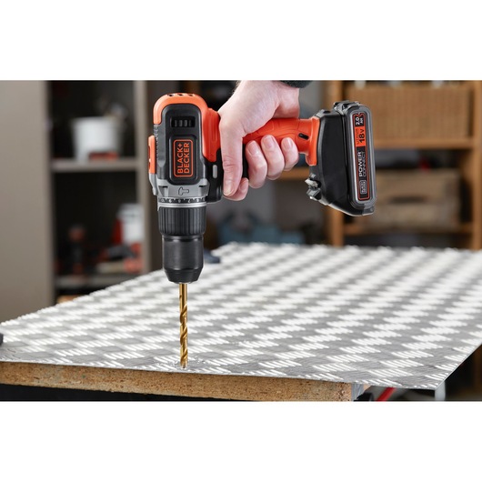 BLACK+DECKER BCD003D2AMB cordless drill in use drilling metal sheet in workshop setting.