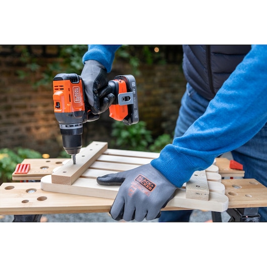 Person using a BLACK+DECKER cordless drill to assemble wooden pieces on a workbench outdoors.