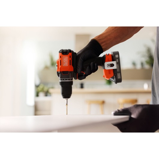 BLACK+DECKER cordless drill in use, drilling a screw into a white surface in a modern indoor setting.