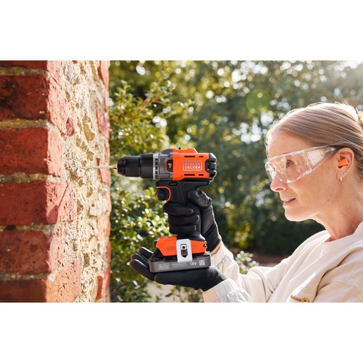 Person drills into outdoor brick wall using a BLACK+DECKER cordless drill, wearing safety glasses and gloves.