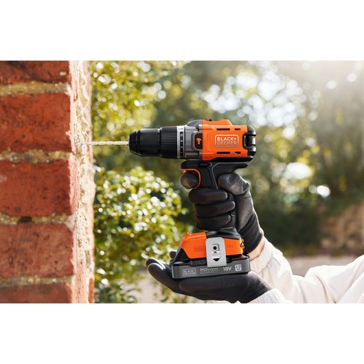 A BLACK+DECKER cordless drill in use, drilling into a brick wall outdoors, held by a gloved hand.