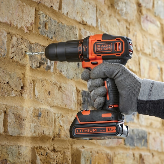 A person wearing grey gloves uses a BLACK+DECKER cordless drill to drill into a brick wall. The drill has an 18V lithium battery.