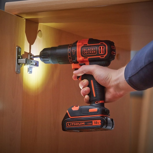 A person is using a BLACK+DECKER cordless drill with an 18V lithium battery to screw or unscrew a metal hinge inside a wooden cabinet, with the drill's built-in light illuminating the work area.