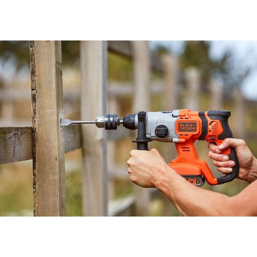 BLACK+DECKER BCD900B rotary hammer drilling into wooden post, orange and black body, close-up outdoor view.