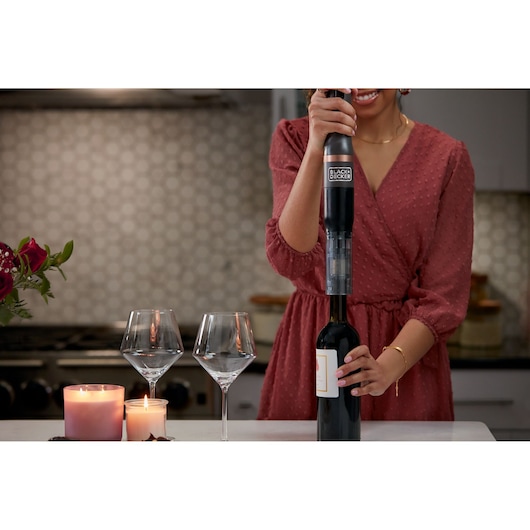 BLACK+DECKER BCKM1011KGF electric wine opener in use opening a wine bottle on a kitchen counter with candles and glasses.