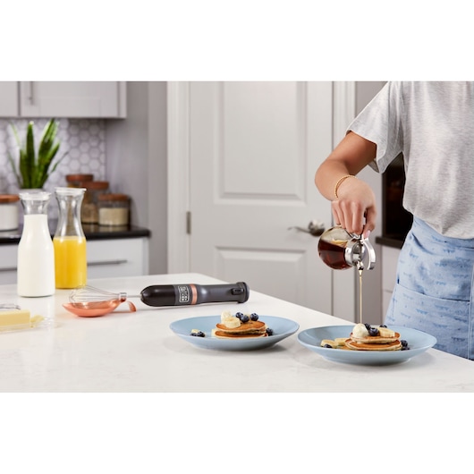 BLACKDECKER BCKM1011KGF hand mixer with whisk on kitchen counter, pancakes with syrup and fruit on blue plates.
