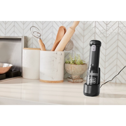 BLACK+DECKER BCKM1012KB black handheld kitchen tool charging on kitchen counter, utensils and plant nearby.