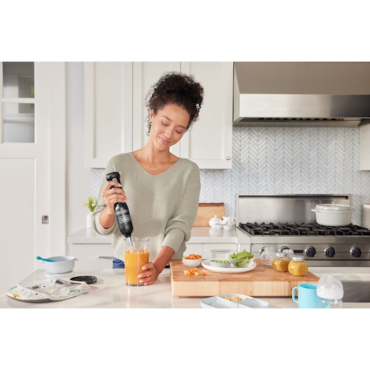 Photo of person using BLACK+DECKER BCKM1016KSB hand blender in a kitchen to blend orange liquid.