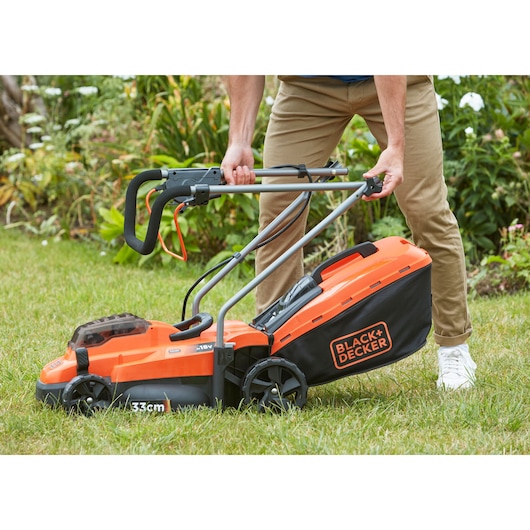 Photo of BLACK+DECKER BCMW3318N orange and black cordless lawn mower being adjusted by a person on grass.