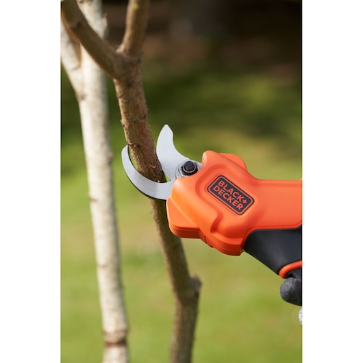 BLACK+DECKER BCPP18D1 orange cordless pruning shears cutting a tree branch outdoors.