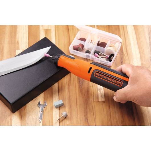 A hand holding a BLACK+DECKER rotary tool is sharpening a large knife blade on a black block. Several tool accessories and attachments are visible, including a wrench, various grinding and sanding bits, and a storage box, all set on a wooden surface.