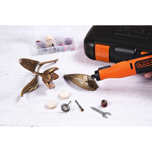 A BLACK+DECKER rotary tool is being used to polish a decorative metal flower. Various rotary tool accessories and a wrench are laid out on the table. In the background, there is a black carrying case and a small organizer box with additional tool bits.
