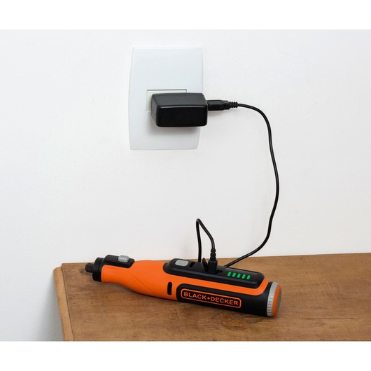 A BLACK+DECKER cordless rotary tool is seen charging on a wooden surface, connected to a wall socket with a black power adapter.