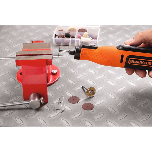 A hand using an orange BLACK+DECKER rotary tool to work on a metal rod held in a red vise. Assorted rotary tool accessories are laid out on the gray patterned surface, including discs, a wire brush, a wrench, and a plastic storage box.
