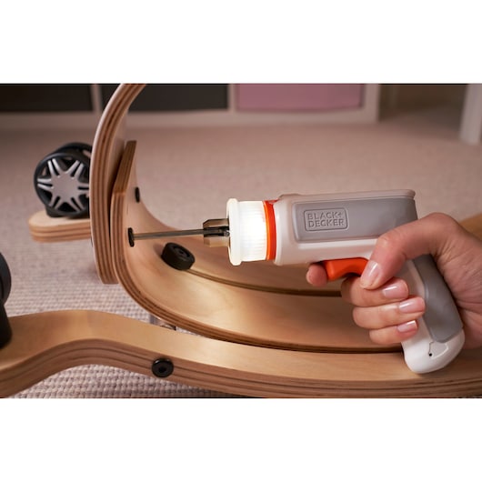 A close-up of a hand using a BLACK+DECKER electric screwdriver to assemble curved wooden pieces with screws. The screwdriver is white and gray with orange accents and is being used to fasten a screw into the wood.
