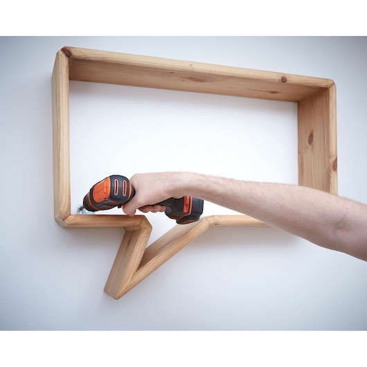 Photo of a BLACKDECKER BDCDD12 cordless drill being used to mount a wooden speech bubble shelf on a wall.