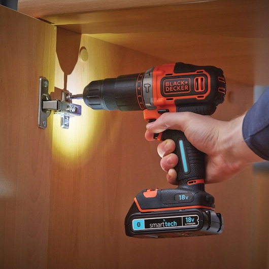 BLACK+DECKER BDCHD18KBST cordless drill driving screw into cabinet hinge, 18V smarttech battery visible.