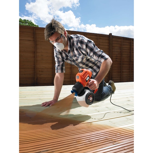Person using BLACKDECKER BDPS200 paint sprayer on outdoor wooden deck, fence in background, low angle.