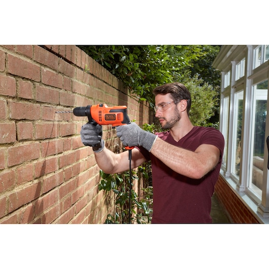Person using a BLACK+DECKER BEH200K corded drill to drill into an outdoor brick wall, wearing gloves.