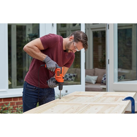 Person uses BLACK+DECKER orange corded drill to make holes in a wooden door outdoors.