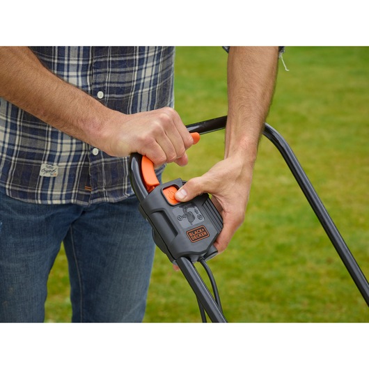 Close-up of hands operating BLACK+DECKER BEMW451 lawn mower handle with control lever on grass.