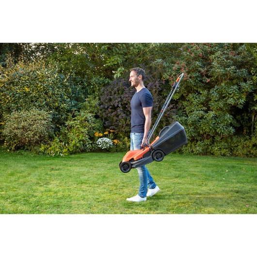 BLACKDECKER BEMW451 lawn mower carried by person in garden with grass and shrubs.