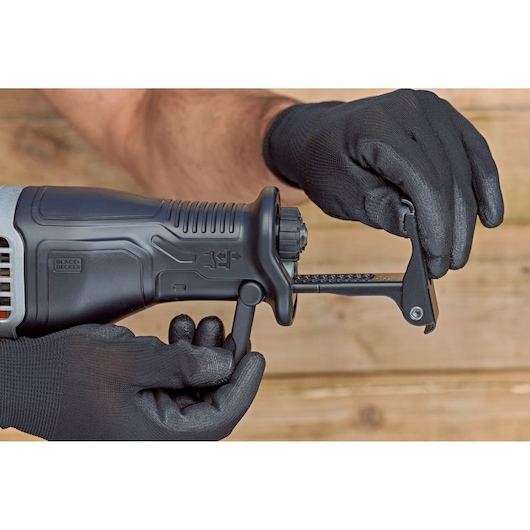 Close-up of gloved hands adjusting the blade clamp on a BLACK+DECKER reciprocating saw, with a wooden background.