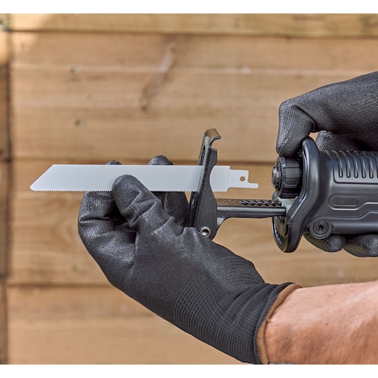 Close-up of a person wearing black gloves installing a blade into a BLACK+DECKER reciprocating saw, with a wooden background.