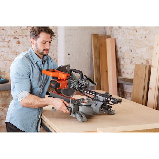 A person using a BLACK+DECKER BES700 sliding mitre saw on a wooden workbench in a workshop. Several wooden boards are stacked against a brick wall in the background.