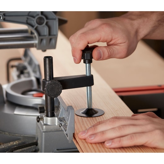 A close-up of hands securing a wooden plank using a clamp on a BLACK+DECKER mitre saw. The clamp holds the wood firmly in place for accurate cutting.