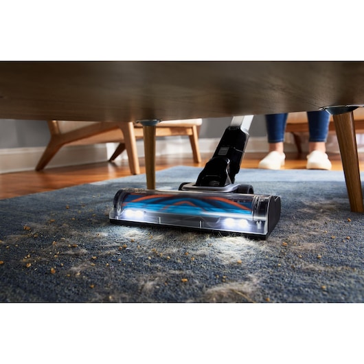 BLACKDECKER BHFEB520D1 stick vacuum cleaning under furniture on blue carpet with LED lights.