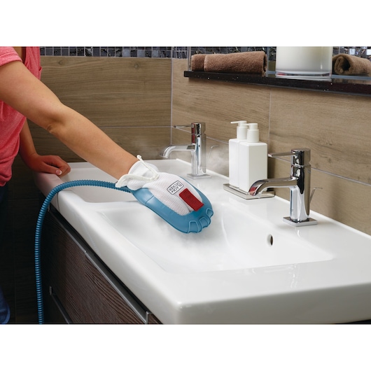 A person cleaning a bathroom sink using a BLACK+DECKER steam cleaning glove. The glove releases steam while wiping the sink surface, and is connected to a hose. Modern faucets and soap dispensers are visible beside the sink.