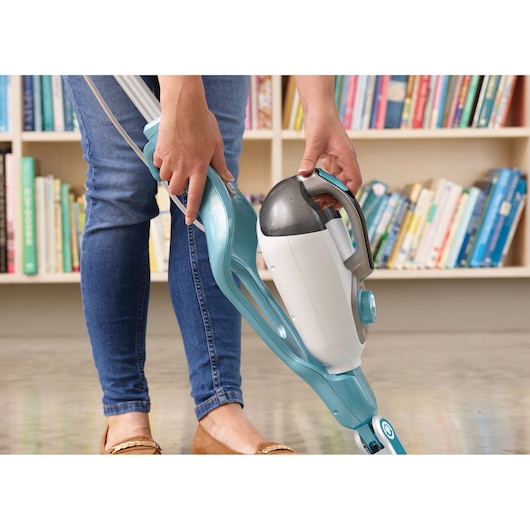 A person is using a BLACK+DECKER steam mop with a detachable handheld unit, standing on a shiny floor in front of a bookshelf filled with books.