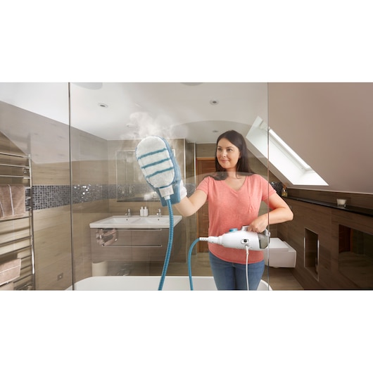A person using a BLACK+DECKER steam cleaning device with a glove attachment to clean a glass shower door in a modern bathroom.