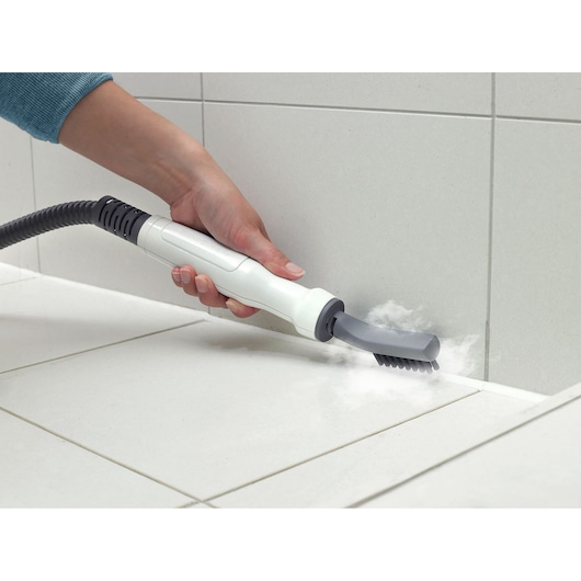 A hand holding a steam cleaning device with a brush attachment, cleaning grout lines between white tiles on a wall and floor.