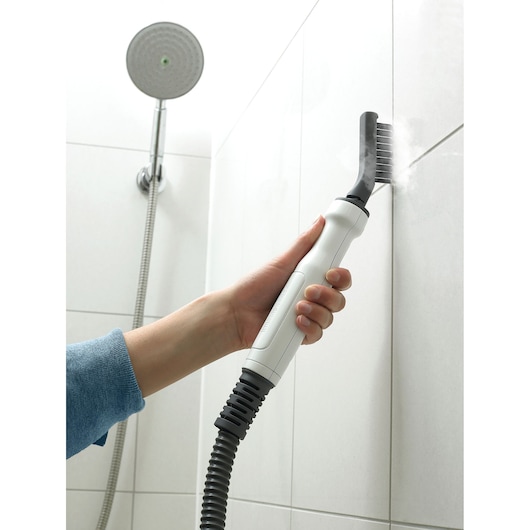 A person uses a BLACK+DECKER steam cleaner with a nozzle attachment to clean the grout lines between white bathroom tiles. A shower head is visible in the background.