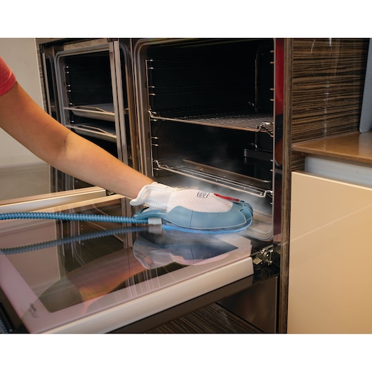 A person is using a BLACK+DECKER steam cleaning glove to clean the inside of an oven. The glove is connected to a hose and releasing steam, effectively removing dirt from the oven door.