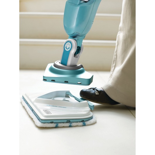 A close-up of a person using their foot to attach a cleaning pad to a blue BLACK+DECKER steam mop, on a tiled floor.