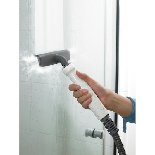 A person uses a handheld steam cleaning device with a grey nozzle to clean a glass shower door, releasing steam to remove grime.