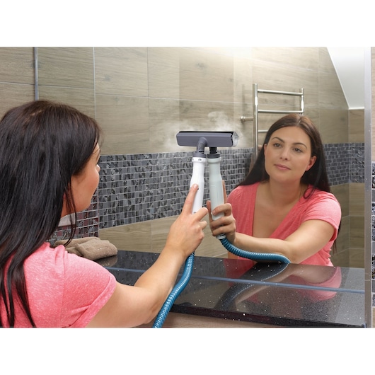 A person in a pink shirt is cleaning a bathroom mirror using a BLACK+DECKER handheld steam cleaner, with steam visible coming from the device.