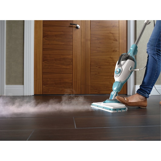A person using a BLUE BLACK+DECKER steam mop to clean a dark tiled floor in front of wooden double doors. Steam is visibly coming from the mop head.