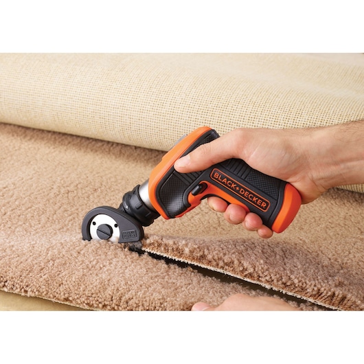 BLACK+DECKER CSCA3 electric rotary cutter trimming beige carpet, close-up of hand holding tool.