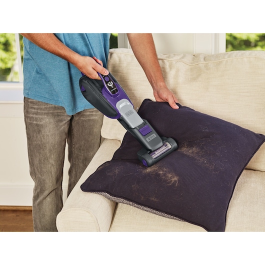BLACKDECKER DVJ325BFSP handheld vacuum cleaning pet hair from a dark cushion on a beige sofa.