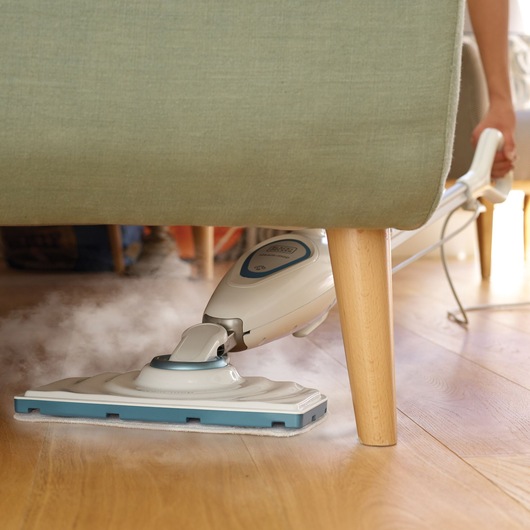 BLACK+DECKER FSM1605 steam mop cleaning wood floor under a green couch, visible steam from mop head.