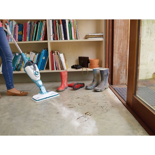 BLACKDECKER FSMH13151SM steam mop cleaning muddy footprints on concrete floor near boots and bookshelf.