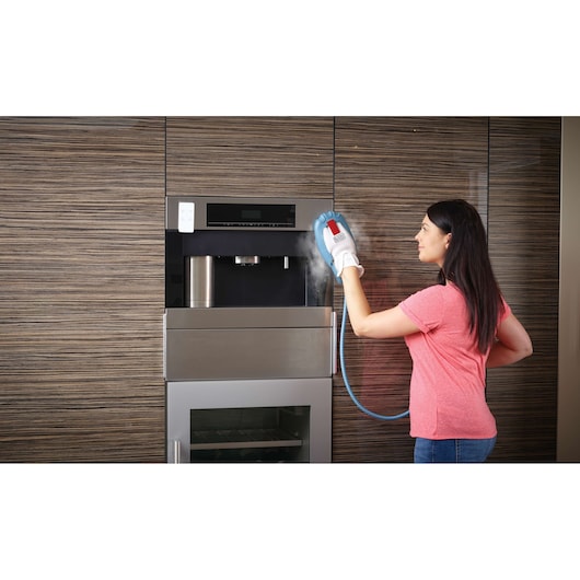A person using a BLACK+DECKER steam cleaning glove to clean a built-in kitchen appliance with wood paneling in the background. Steam is visible coming from the glove.