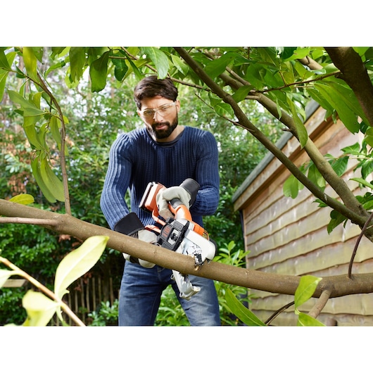 A person with a blurred face is using a BLACK+DECKER battery-powered chainsaw to cut a tree branch in a garden, wearing gloves and a blue sweater. There are green leaves and a wooden shed in the background.
